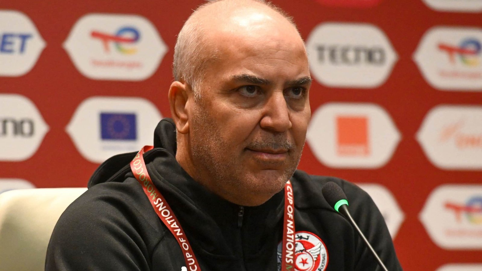 Tunisia's national team coach, focused and serious, speaks at a press conference backdrop featuring sponsorship logos and national colours.