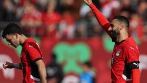 Egyptian players celebrate during a match, wearing red kits, with one player raising his arm in triumph while another looks down.