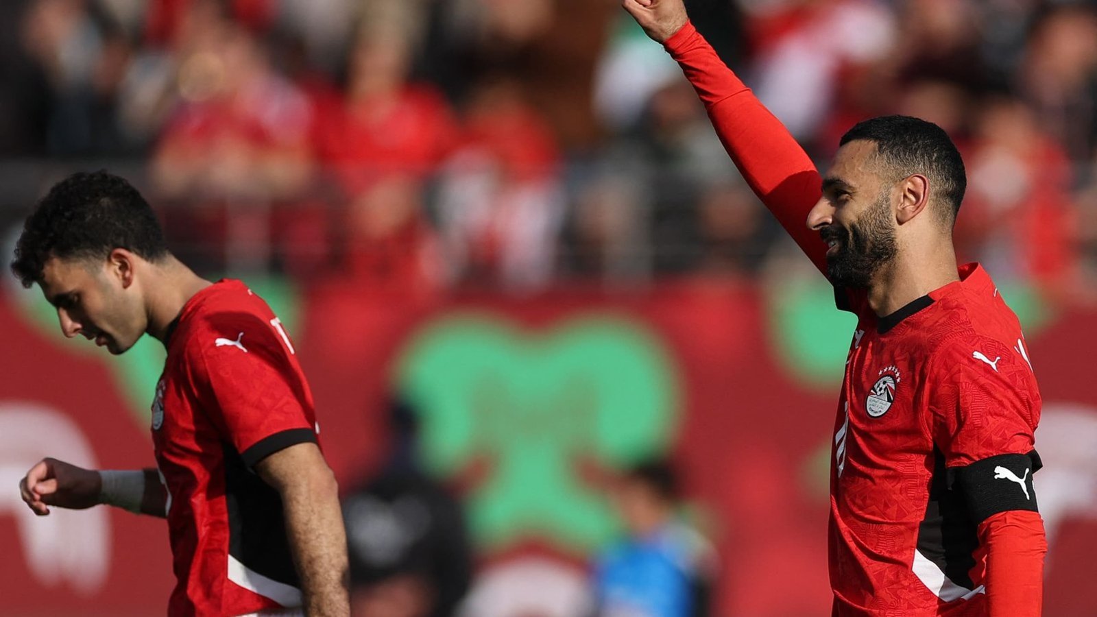 Egyptian players celebrate during a match, wearing red kits, with one player raising his arm in triumph while another looks down.