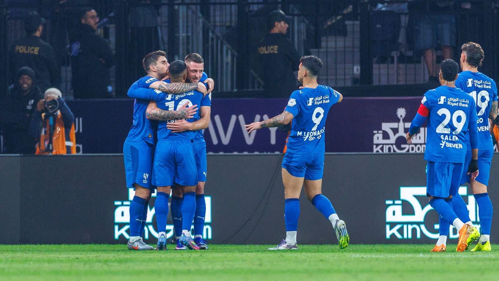 Players in blue jerseys celebrate a goal, embracing each other on the pitch. Notable numbers include 9 and 29, with a stadium backdrop.