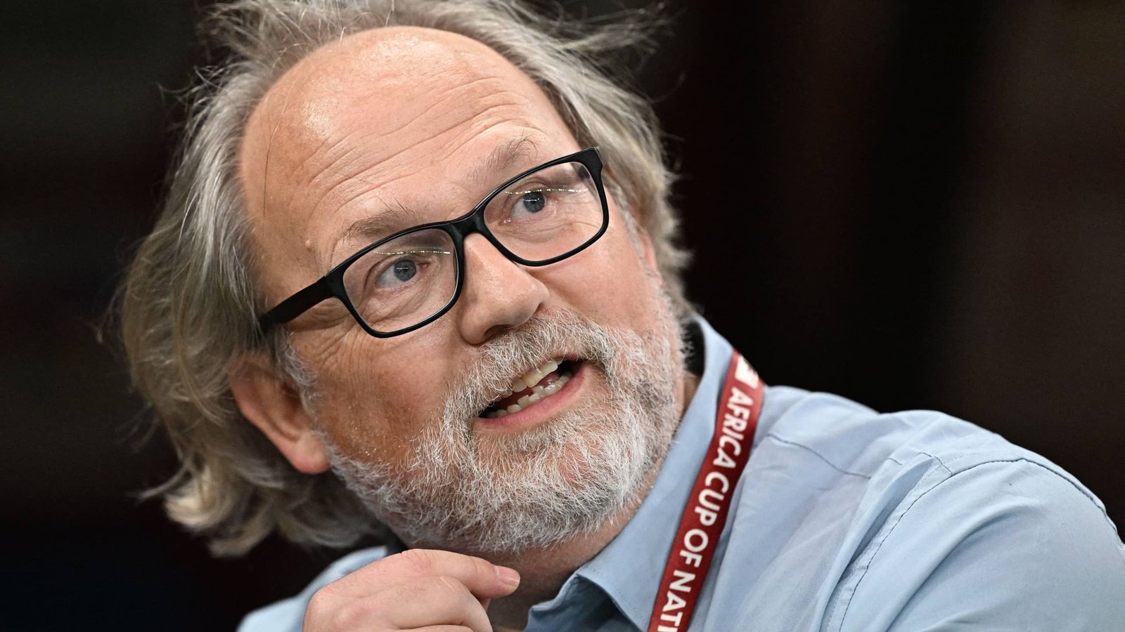 A man with glasses and dishevelled hair gestures while discussing the Africa Cup of Nations, wearing a lanyard for the event.