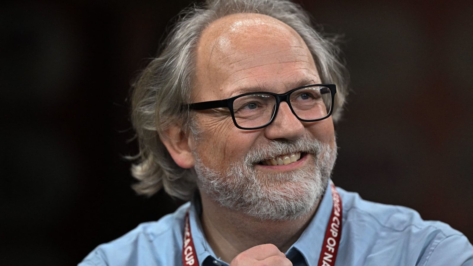 Commentateur sportif souriant, portant des lunettes et ayant des cheveux gris, avec un lanyard, assis dans un stade, probablement lors d'un événement footballistique.