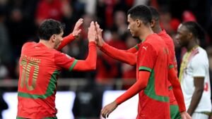 Moroccan players celebrate a goal, wearing red jerseys with green accents, during an international match against Spain.