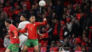 Morocco players number 9 and 23 challenge for the ball against an opposing player, with a sea of fans in red cheering in the background.