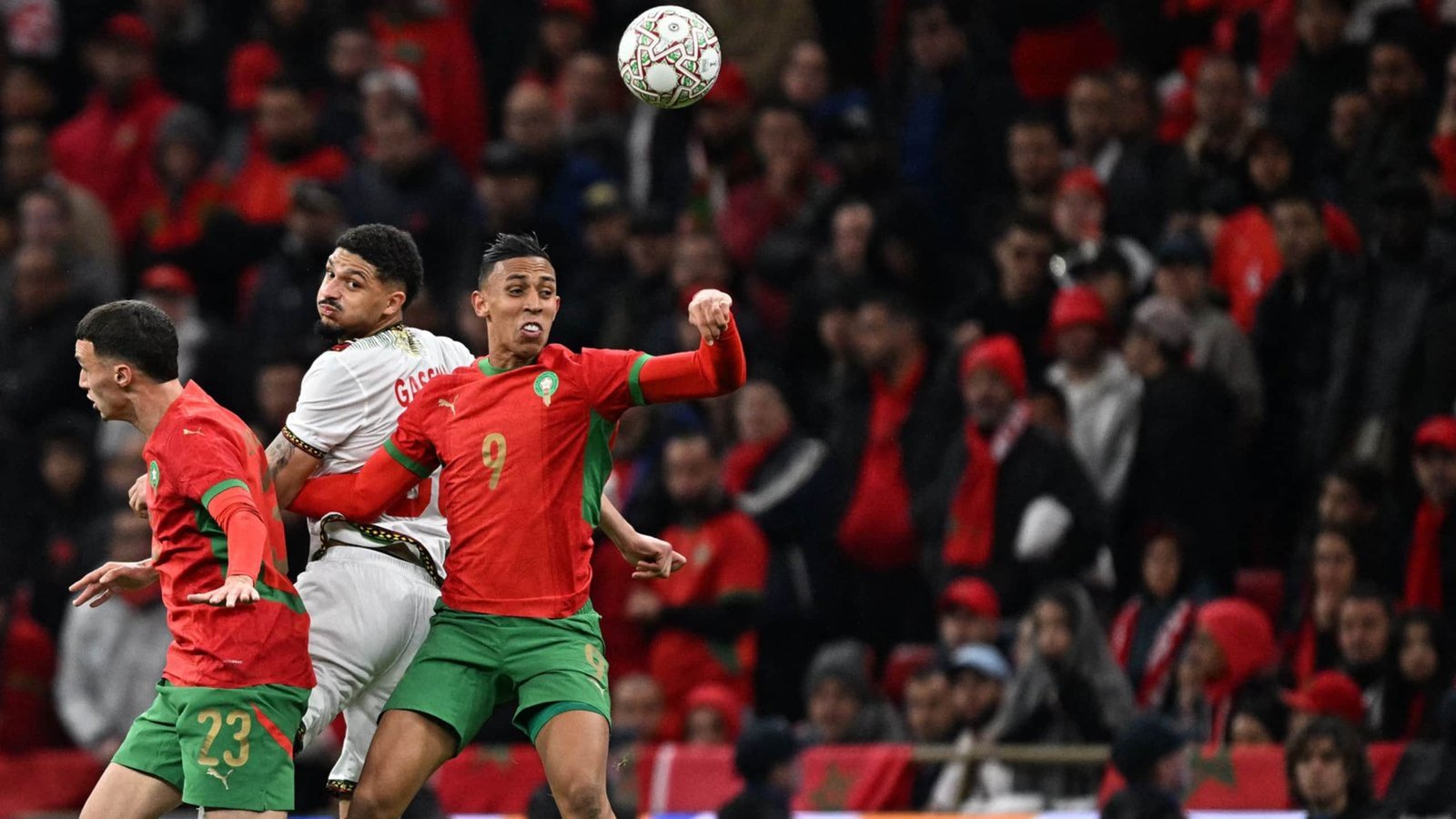 Morocco players number 9 and 23 challenge for the ball against an opposing player, with a sea of fans in red cheering in the background.