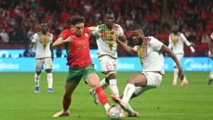 Moroccan midfielder in a red and green kit dribbles past a defender in white during a tense match against Mali, with fans in the backdrop.