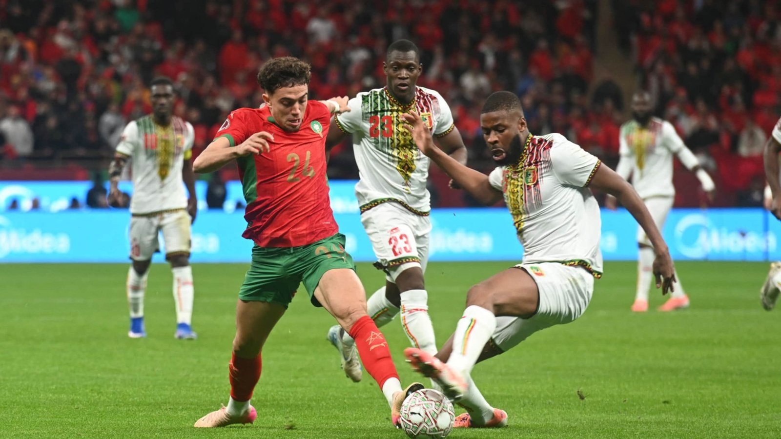 Moroccan midfielder in a red and green kit dribbles past a defender in white during a tense match against Mali, with fans in the backdrop.