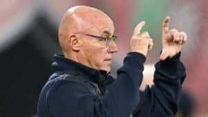 Coach gesturing tactically from the sidelines during a match, wearing a dark jacket and glasses, with a blurred stadium background.