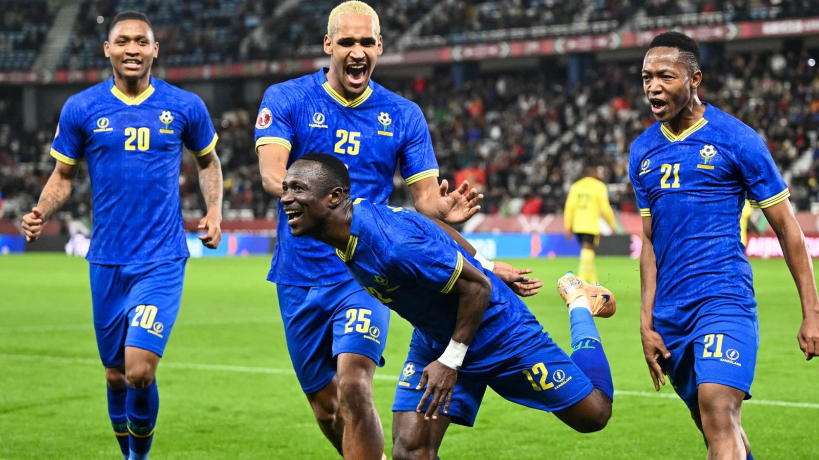 Celebrating players in blue jerseys, including number 25, joyfully reacting after scoring a goal, with excited teammates nearby.