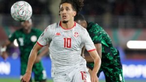 Tunisia's number 10 player, in a white kit with red accents, chases the ball during a match against Nigeria, wearing a determined expression.