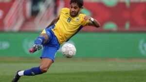 Een speler in een geel-blauw tenue probeert een schot tijdens een wedstrijd, met een vervaagde bal in de lucht op het groene veld.