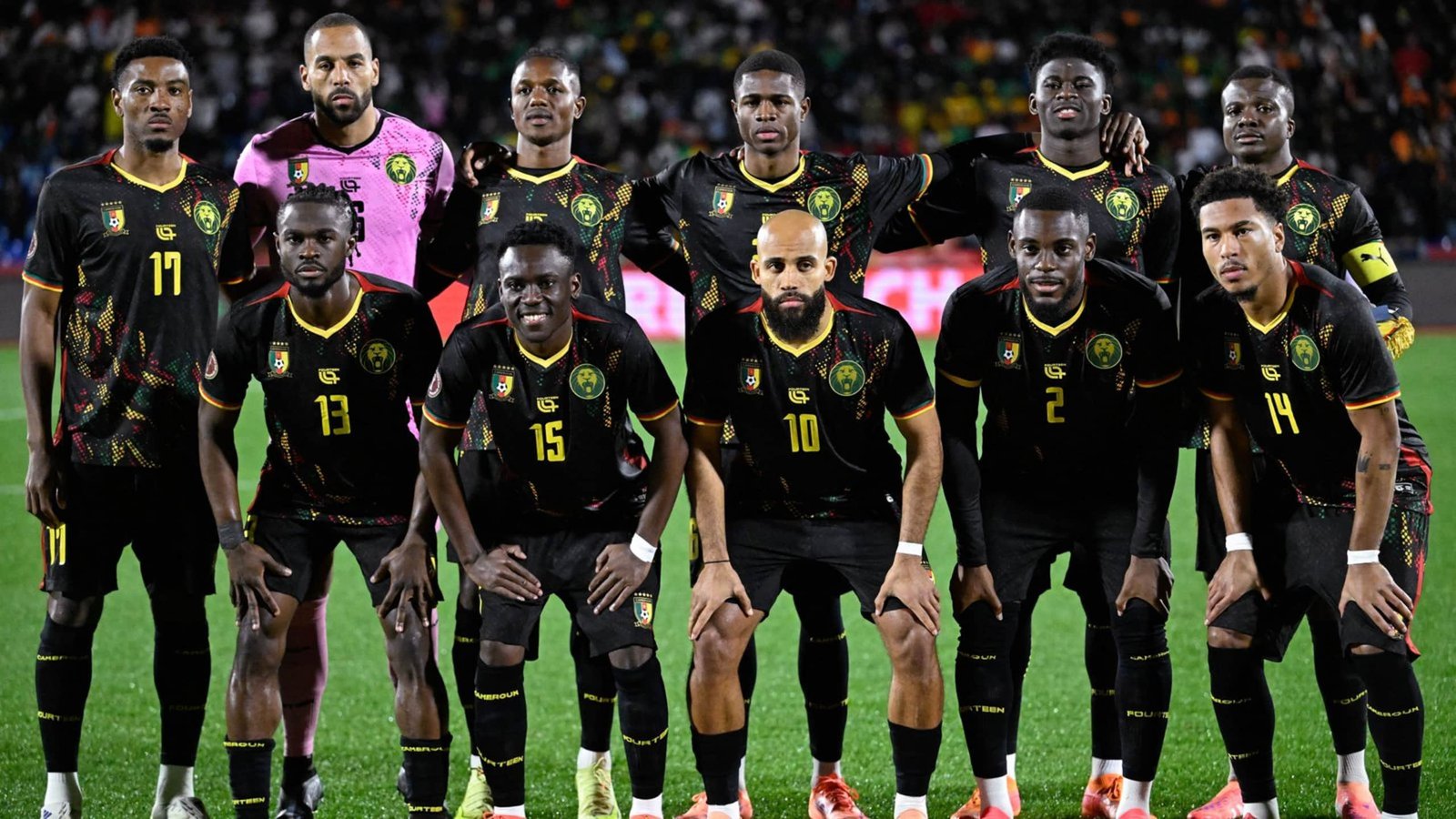 Cameroon national team lineup featuring players in black and yellow kits, posing on the pitch with vibrant stadium backdrop.
