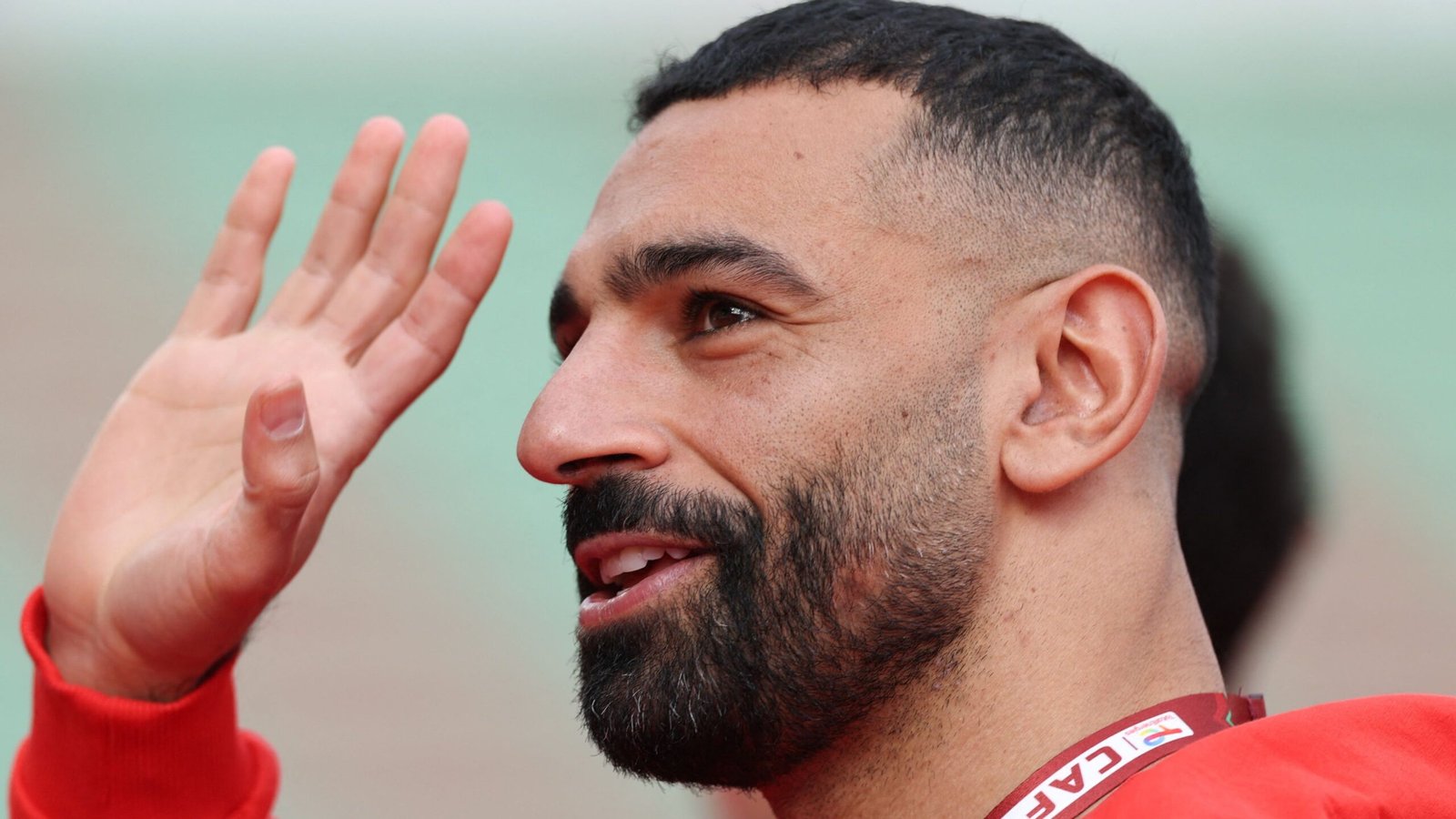 Moroccan football player waves, smiling, wearing a red training kit with a visible badge, against a blurred background.