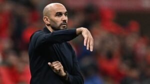 Walid Regragui gestures on the sideline in a black shirt, with a blurred background of fans in red, during a match involving Morocco.