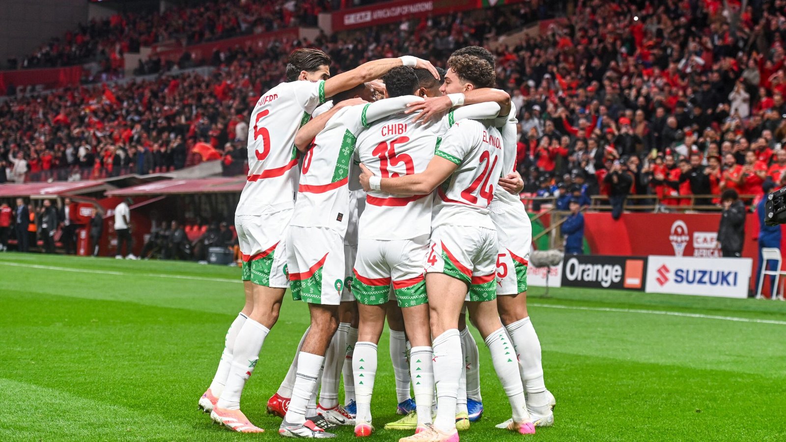 Marokkaanse spelers vieren een doelpunt, omhelzend op het veld met juichende fans op de achtergrond, die levendige teamtenues en een feestelijke sfeer tonen.