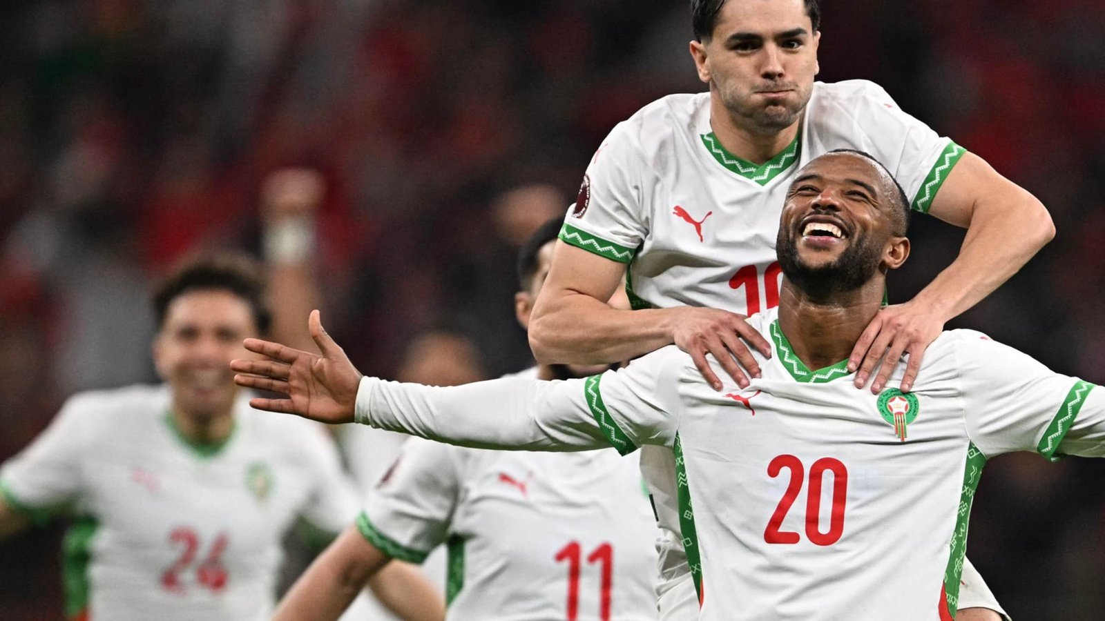 Moroccan players celebrate joyfully after a goal, wearing white kits with green accents, amidst a lively stadium atmosphere.