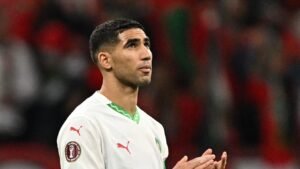 Achraf Hakimi in Morocco's national team kit, looking upwards in a moment of reflection during a match, with a blurred stadium background.