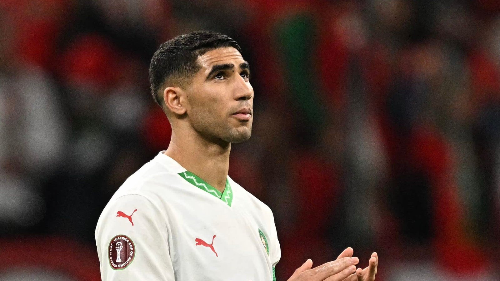 Achraf Hakimi in Morocco's national team kit, looking upwards in a moment of reflection during a match, with a blurred stadium background.
