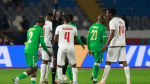 Senegal and Benin players discuss a foul during a tense moment in a match, featuring green and white team kits with vibrant stadium backdrop.