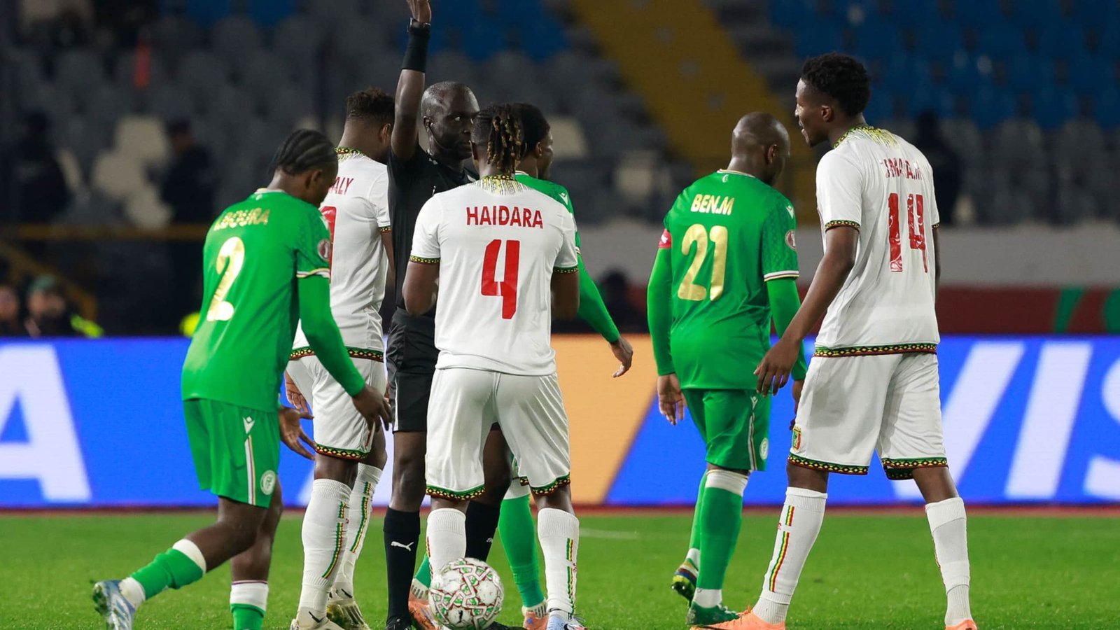 Senegal and Benin players discuss a foul during a tense moment in a match, featuring green and white team kits with vibrant stadium backdrop.