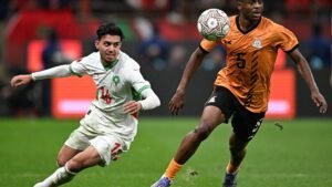 Moroccan player wearing white, number 16, lunges for the ball against a Zambian opponent in orange, number 5, during an intense match action.