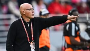 Coach giving tactical instructions during an Africa Cup of Nations match, wearing a black sweater and accreditation lanyard, with focused intensity.