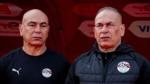 Des entraîneurs de football égyptiens en vestes d'équipe, arborant les écussons de l'équipe nationale, dans un banc de touche à thème rouge avant un match.