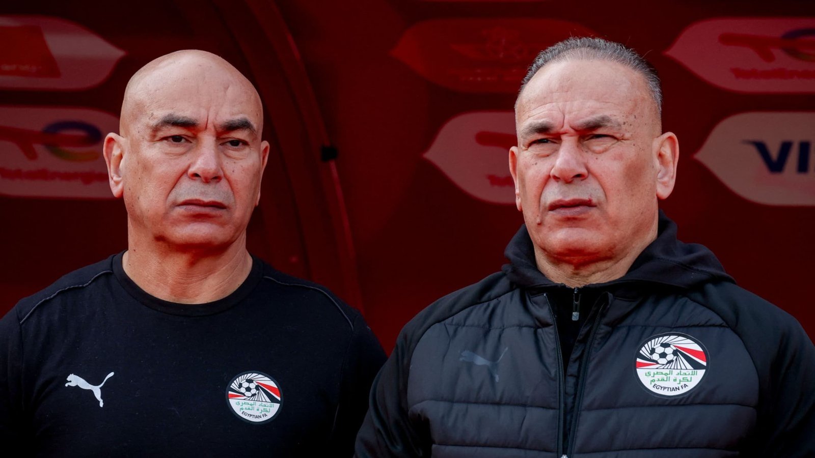 Des entraîneurs égyptiens vêtus de tenues de l'équipe nationale, se tenant sérieux devant un fond rouge, reflétant des préparatifs de match tendus.