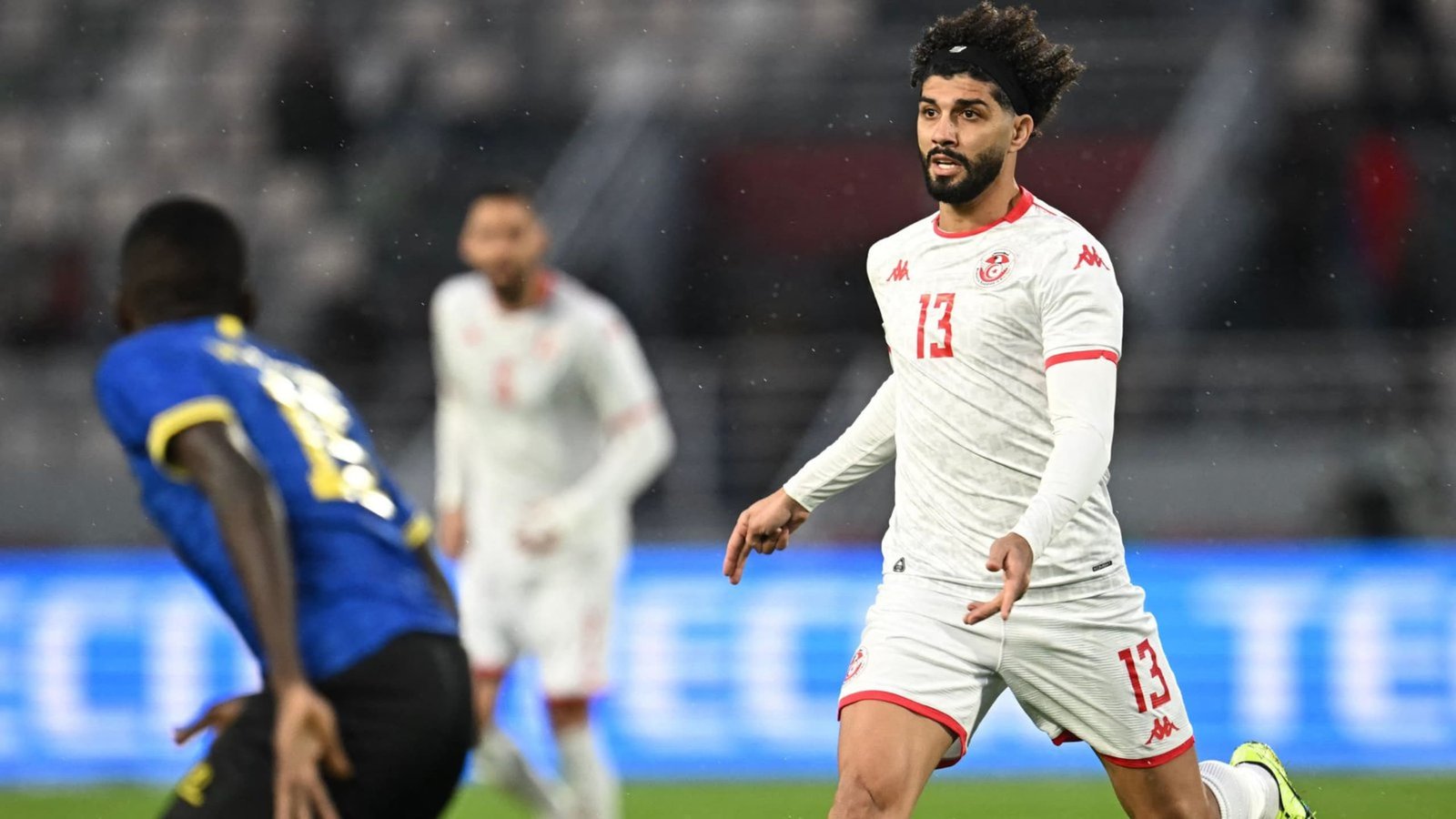 Tunisia's player, wearing white with red accents and number 13, sprinting towards the ball as an opponent in blue defends during a match.