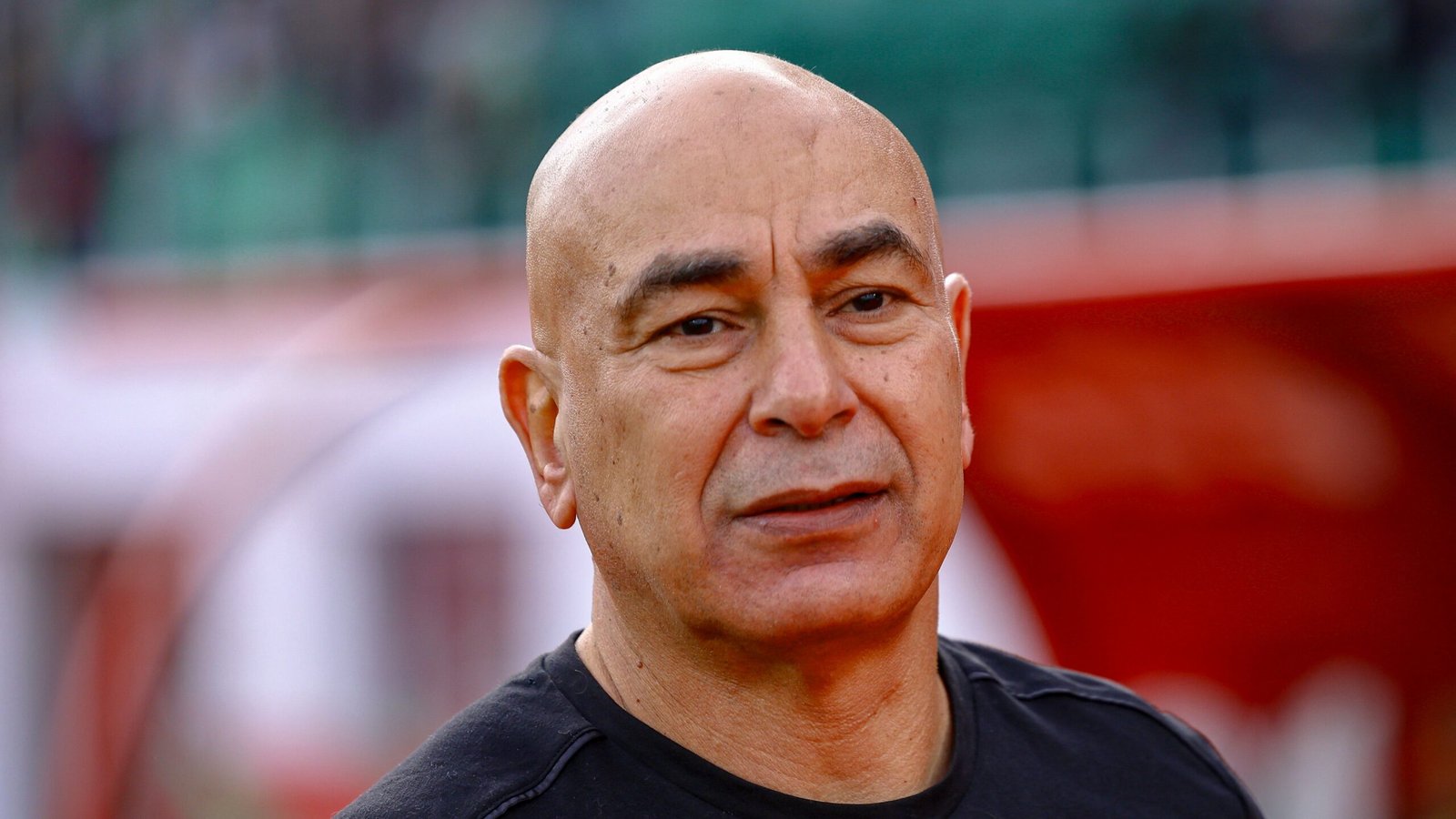 Moroccan football coach with a bald head, wearing a black shirt, stands against a blurred stadium backdrop, reflecting concentration.