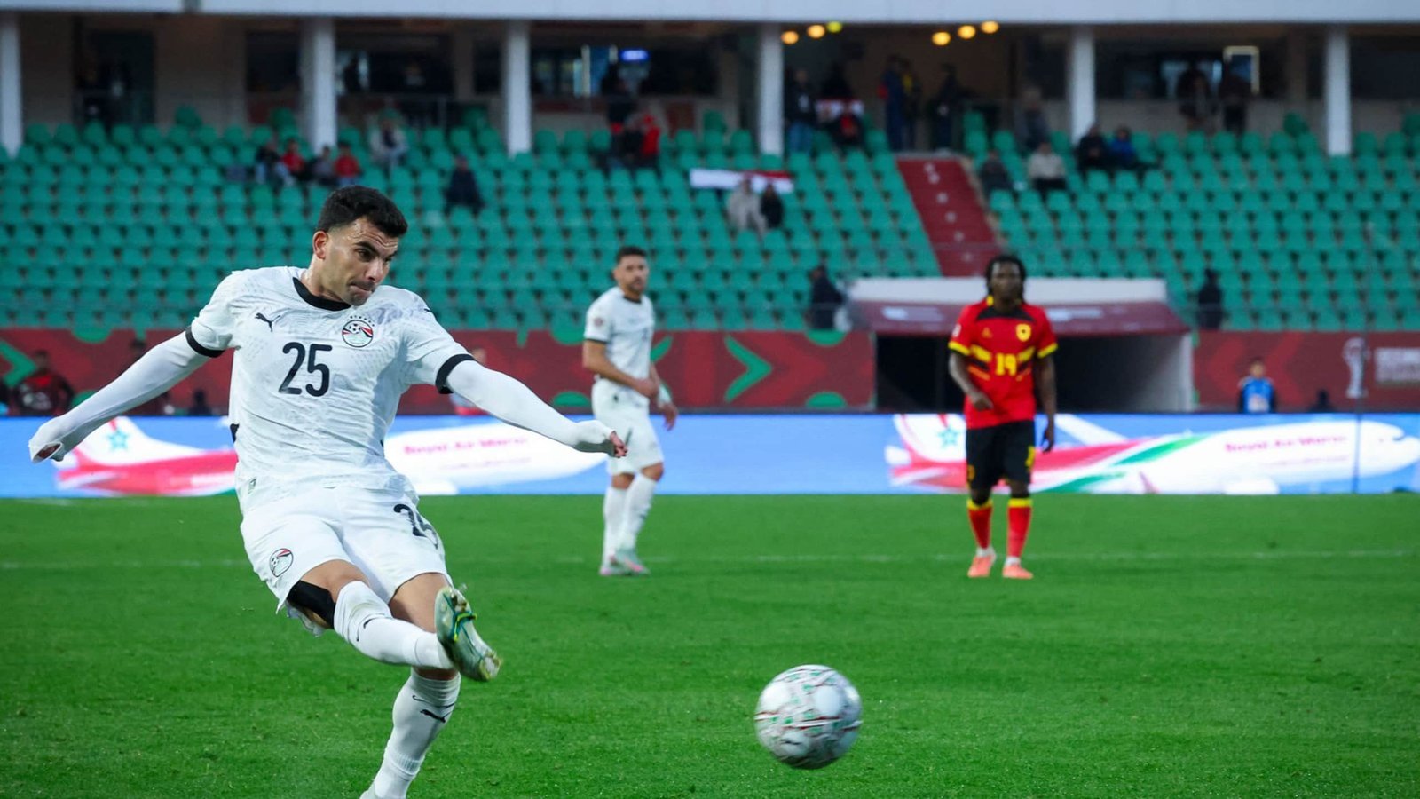 Egyptian player wearing jersey number 25 striking a penalty kick against Angola, with green spectators' seating in the background.