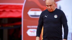 Egyptian coach in a black Puma kit, looking pensive as he walks past a backdrop featuring Egypt's flag during training.