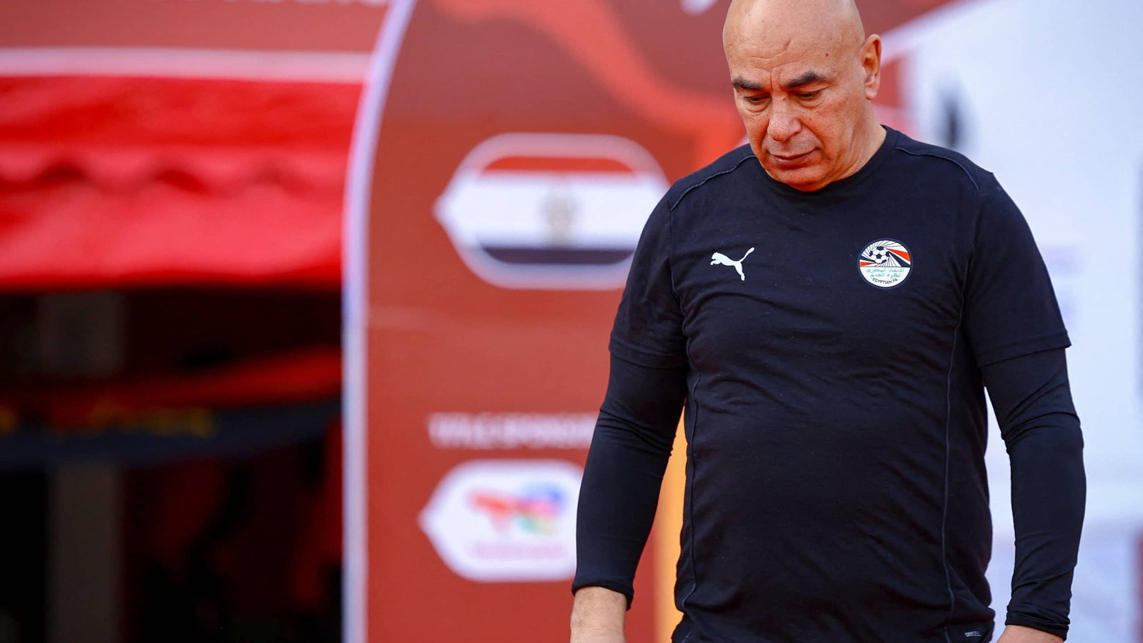 Egyptian coach in a black Puma kit, looking pensive as he walks past a backdrop featuring Egypt's flag during training.