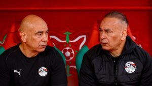 Deux entraîneurs de football, vêtus de noir, sont assis sur le banc en pleine discussion, devant un fond rouge vif orné de motifs marocains.
