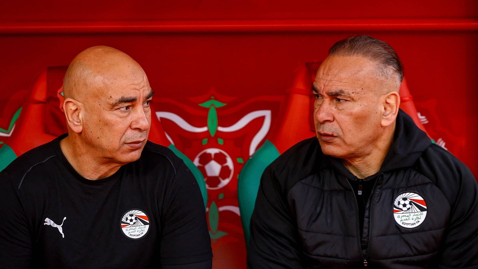 Two football coaches sit side by side on a bench, wearing matching black training kits with Egyptian Football Association logos, against a red backdrop.