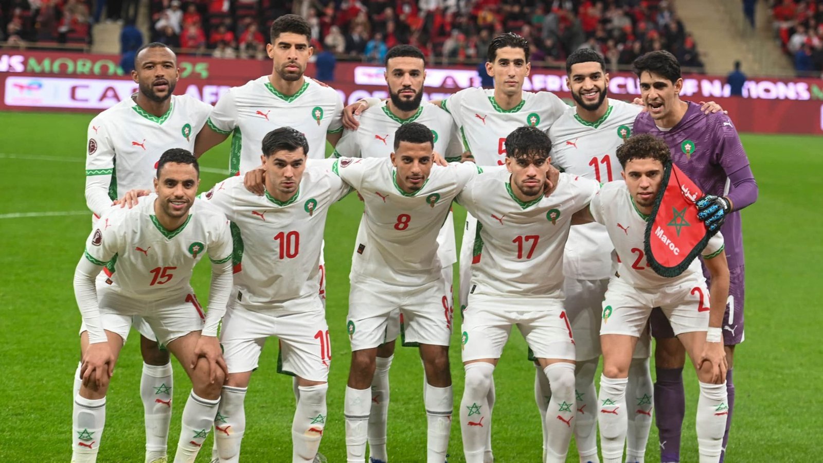 Morocco's national football team posing in white kits with green accents, celebrating their participation in a major tournament, with a vibrant crowd in the background.