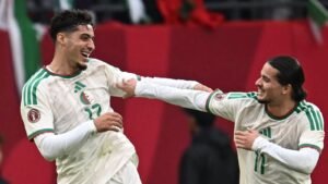 Algerijnse spelers die vreugdevol feestvieren tijdens een wedstrijd, gekleed in witte tenues met groene accenten, tegen een levendige rode achtergrond.