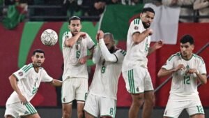 Des joueurs algériens en tenue blanche rehaussée de vert forment un mur alors qu'un ballon s'apprête à être frappé lors d'une situation de match tendue.
