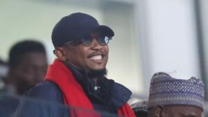 Smiling fan wearing a navy cap and sunglasses with a red scarf, enjoying the match atmosphere, alongside another supporter in traditional attire.