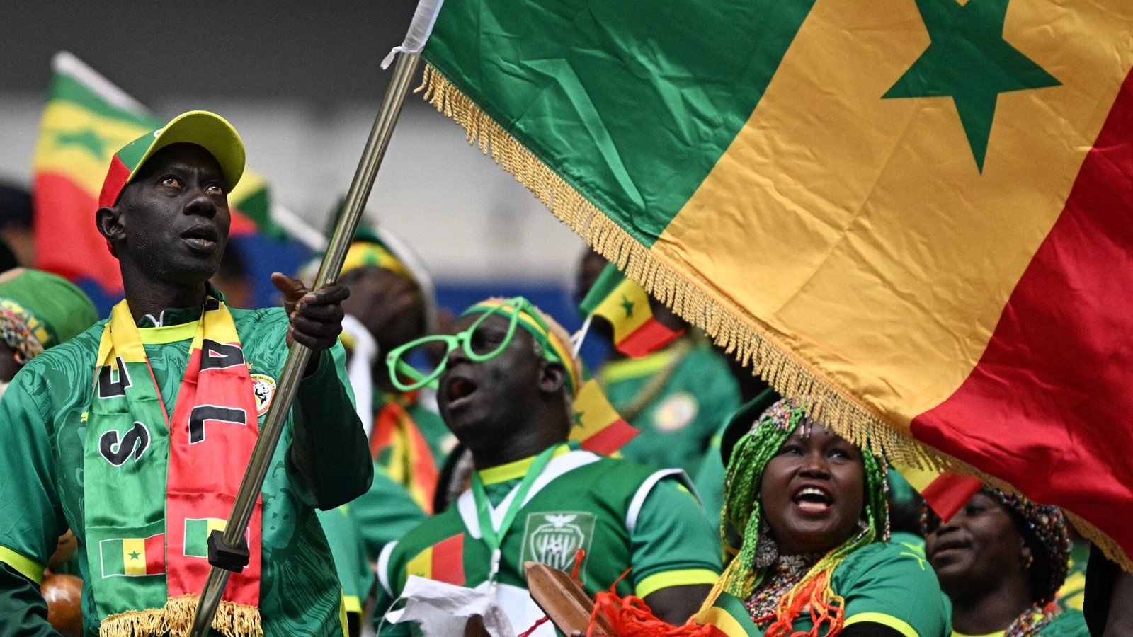 Les fans sénégalais célèbrent passionnément, agitant un grand drapeau aux couleurs vives vert, jaune et rouge, portant les couleurs et accessoires de l'équipe.