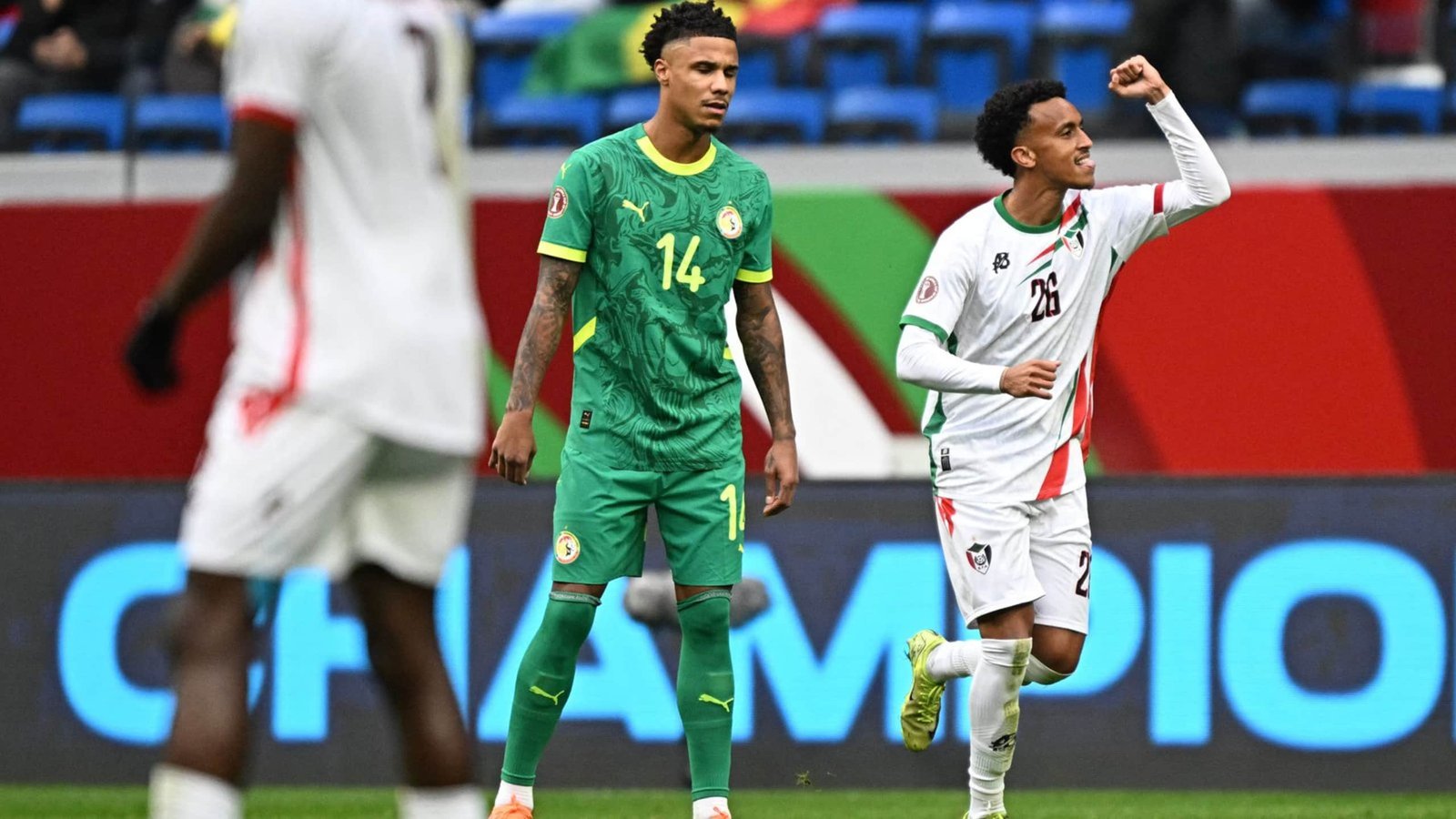 Moroccan player celebrating a goal with a clenched fist, while Senegalese player, in green kit, looks on in a tense moment of the match.