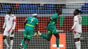 Senegalese players celebrate a goal, with one in a green kit and another in a blue kit, while opponents look on in a tense moment during the match.