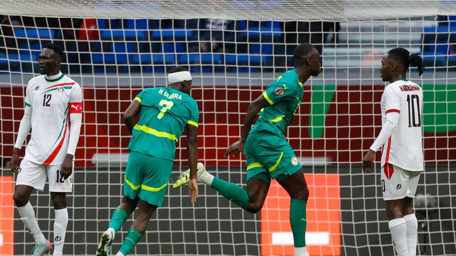 Senegalese players celebrate a goal, with one in a green kit and another in a blue kit, while opponents look on in a tense moment during the match.