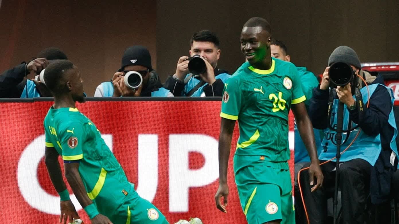 Senegalese spelers vieren een doelpunt, dragen groene tenues met gele accenten, terwijl fotografen het moment vastleggen op de achtergrond.