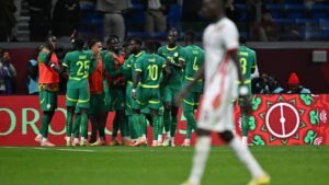 Senegal team celebrates a goal, wearing green kits with yellow accents, while fans cheer in the background during a match.