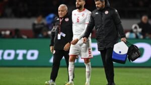 Tunisia's number 6 player is assisted off the pitch by coaching staff, showcasing concern and determination during a match.