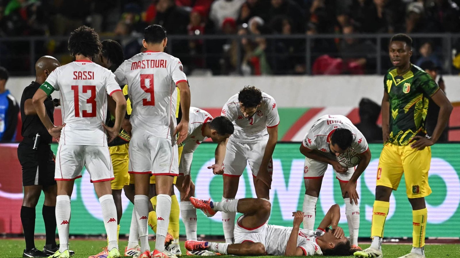 Tunisian players Sassi and Mastouri gather around an injured teammate during a match against Mali, with concerned expressions and a referee present.