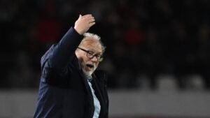 Coach gebaart gepassioneerd tijdens een wedstrijd, draagt een bril en een donker jasje, en toont intense emotie aan de zijlijn.