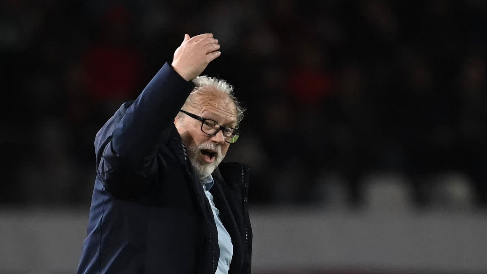 Coach gebaart gepassioneerd tijdens een wedstrijd, draagt een bril en een donker jasje, en toont intense emotie aan de zijlijn.