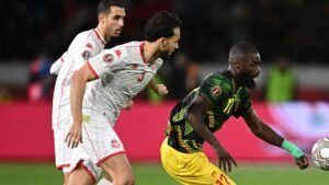 Tunisia's defenders challenge a Gambian forward during an intense match, showcasing the vibrant colours of team kits and a lively stadium atmosphere.